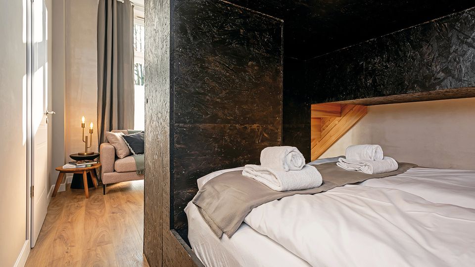 Modernes Schlafzimmer mit großem Bett, weißer Bettwäsche und Handtüchern, eingerahmt von einer dunklen, strukturierten Wandverkleidung und einem Holzelement am Kopfteil. Daneben ein kleiner Wohnbereich mit Sofa, Beistelltisch und Lampe sowie hohen Fenstern mit Vorhängen und hellem Holzboden.