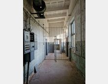 Ein leerer, verfallener Raum mit weiß gestrichenen Ziegelwänden, einem offenen Gittertor in der Mitte und sichtbaren Kabeln sowie alten Schalttafeln an der linken Wand. Rechts befinden sich Fenster mit bröckelndem Putz, und die Decke zeigt eine Betonstruktur mit quadratischen Vertiefungen.