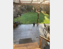 Eine überdachte Terrasse mit Fliesenboden und Holzstufen, die in einen Garten führt. Der Garten hat eine Rasenfläche, umgeben von Büschen und kleinen Dekorationen, wie hängende Ornamente und Blumentöpfe.