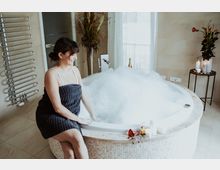 Eine Frau sitzt in einem Badezimmer am Rand einer runden Whirlpool-Badewanne mit Schaum, während Kerzen, Rosenblätter und kleine Flaschen auf dem Rand dekoriert sind. Im Hintergrund befindet sich ein Handtuchwärmer, große Fenster und Blumenarrangements.