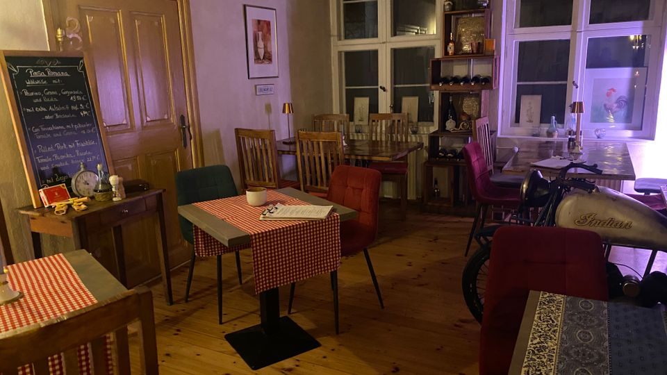 Gemütlicher Gastraum eines kleinen Restaurants mit Holzboden, karierten Tischdecken, bunten Stühlen und einer Kreidetafel mit Speisekarte neben einer Holztür. Im Hintergrund stehen hohe Fenster, ein Regal mit Weinflaschen und ein Thekentisch; rechts ist ein dekoratives Motorrad im Innenraum zu sehen.