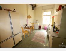 Ein einfacher Küchenraum mit cremefarbenen Schränken, einer kleinen Sitzecke mit Holztisch und gelben Stühlen sowie einer Arbeitszeile. Der Boden ist mit einem grünen Belag und einem bunten Teppich ausgelegt, und an der Decke sind sichtbare Beschädigungen erkennbar.