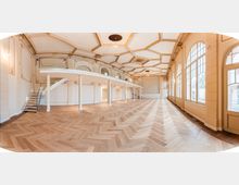 Großer, leerer Saal mit hellem Fischgrät-Parkett, hoher kassettierter Decke mit Stuck und umlaufender Empore mit Treppen. Rechts sorgen bodentiefe, bogenförmige Fensterfronten und eine Glastür für viel Tageslicht.