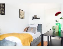 Modern eingerichtetes Schlafzimmer mit einem Einzelbett, das mit grauem Stoff gepolstert ist, weißen Kissen und einer gelben Decke. An der Wand hängen zwei Bilder, ein Regal mit Dekorationen, und im Vordergrund steht ein schwarzer Tisch mit grünen Vasen und einer roten Kunstblume.