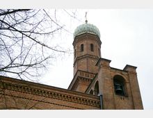 Backstein-Gebäude mit einem quadratischen Glockenturm und einer Kuppel, umgeben von kahlen Baumästen vor einem bewölkten Himmel. Die Architektur zeigt dekorative Backsteinmuster und Rundbogenfenster.