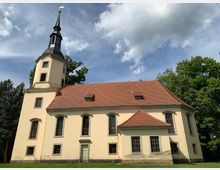 Historische Kirche mit beigefarbener Fassade und rotem Ziegeldach, flankiert von mehreren großen Fenstern mit schwarzen Gittern. Der Kirchturm hat eine dunkle Spitze und ist mit einer Wetterfahne versehen. Im Hintergrund sind Bäume und ein blauer Himmel mit weißen Wolken zu sehen.