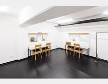Ein heller Schminkraum mit weiß gestrichenen Wänden, schwarzem Boden und zwei Schminktischen, jeweils mit zwei Holzstühlen und integrierten Spiegeln. An der Decke ist eine sichtbare schwarze Rohrleitung angebracht, daneben steht ein leerer Kleiderständer auf Rollen.