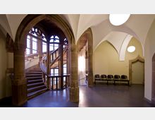 Innenraum eines historischen Gebäudes mit einer gewundenen Holztreppe, großen gotischen Fenstern mit Sprossen und einem Gewölbebogen. Auf der rechten Seite stehen mehrere schwarze Stühle vor einer hellgelben Wand mit einer Tür.