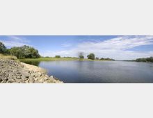 Flusslandschaft mit klarem Wasser, grasbewachsenen Ufern und vereinzelt stehenden Bäumen; im Vordergrund ist das Ufer mit Steinen und Sand bedeckt, der Himmel ist teilweise bewölkt.