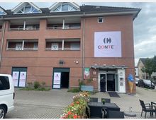 Backstein-Gebäude mit drei Etagen und einem italienischen Restaurant im Erdgeschoss namens „Conte's Pizza & Pasta“. Vor dem Eingang stehen eine italienische Flagge, ein großes Kunstwerk in Form einer Eiswaffel und Sitzgelegenheiten auf einer gepflasterten Terrasse mit Blumenkästen.