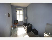 Schlicht eingerichtetes Zimmer mit blauen Wänden und einem einzelnen Fenster. Im Raum befinden sich ein kleines Sofa aus schwarzem Leder, ein alter Röhrenfernseher auf einem runden Holztisch, ein Glas-Beistelltisch mit einer Pflanze und eine Stehlampe.