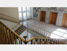 Offenes Treppenhaus in einem Gebäude mit gold-schwarzen Geländern, hellen Fliesenböden und großen Fenstern, die Tageslicht hereinlassen. Im Hintergrund ist eine Wand mit Mosaikkunst und drei Holztüren sichtbar.