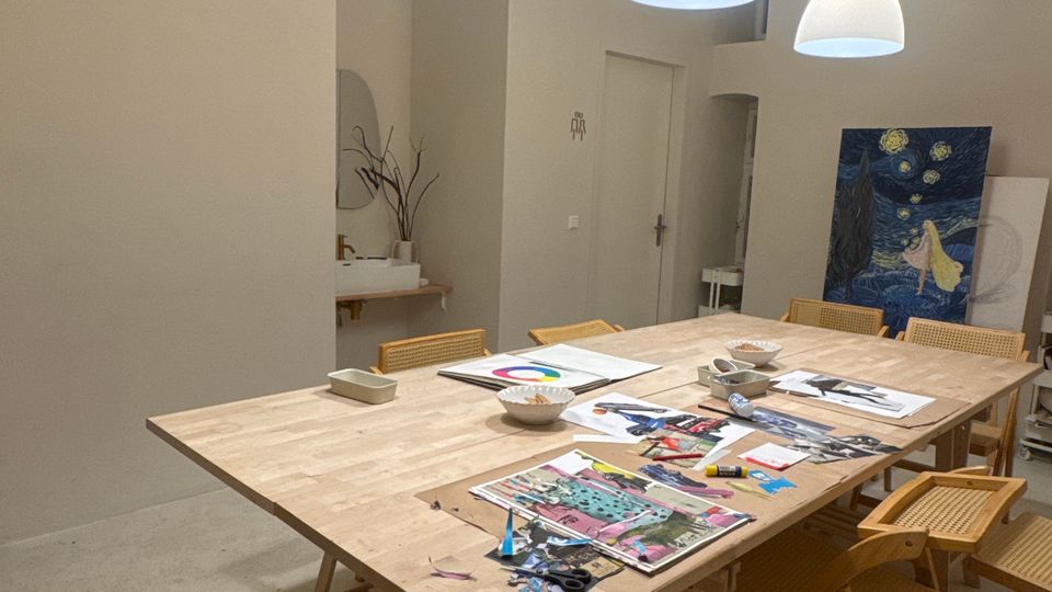 Heller Atelierraum mit großem Holzarbeitstisch und mehreren Holzstühlen, auf dem Tisch liegen Papier, Scheren und Bastelmaterialien. Von der hohen Decke hängen drei weiße Pendelleuchten; im Hintergrund stehen eine kleine Ablage und ein großes Bild an der Wand.