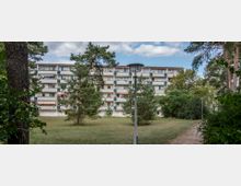 Ein mehrstöckiges Wohngebäude mit Balkonen, umgeben von Grünflächen, Kiefern und Sträuchern. Im Vordergrund befindet sich ein Gartenweg mit Laternen, und der Himmel ist teilweise bewölkt.