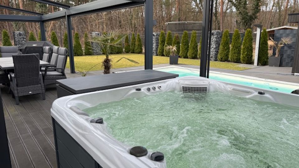 Outdoor patio with a large white hot tub on a dark deck beneath a modern metal pergola, with a dining table and chairs nearby. In the background are a lawn bordered by tall shrubs and stone privacy walls, with a wooded area beyond.