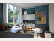 Eine moderne Wohnküche mit weißer Einrichtung, einer großen Fensterfront, blau gestrichenen Wänden und hängenden Deckenleuchten. Auf der Küchenarbeitsfläche befinden sich Pflanzen, Geschirr und eine Obstschale, und im Hintergrund steht ein Holzbank-Sitzbereich neben einem bunten Gemälde.