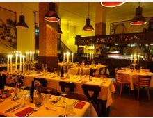 Ein stilvoll eingerichtetes Restaurant mit gedeckten Tischen, weißen Tischdecken und silbernen Kerzenhaltern mit brennenden Kerzen. Der Innenraum ist warm beleuchtet, mit industriellen Hängelampen, Mosaiksäulen und dekorativen Wandbildern im Hintergrund.