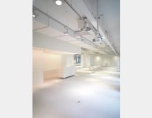 Heller, moderner Innenraum mit minimalistischem Design, weißem Boden und Decke, ausgestattet mit schlichten Sitzreihen und einem weißen Empfangstresen. Die Decke weist sichtbare Beleuchtungsschienen, Lüftungsauslässe und Projektoren auf.