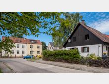 Wohnsiedlung mit mehreren freistehenden Häusern, darunter ein weißes Haus mit dunkler Holzfassade und ein gelb-weißes Mehrfamilienhaus. Im Vordergrund eine Straße mit Gehweg, gesäumt von Bäumen und kleinen Gärten mit Holzzäunen.