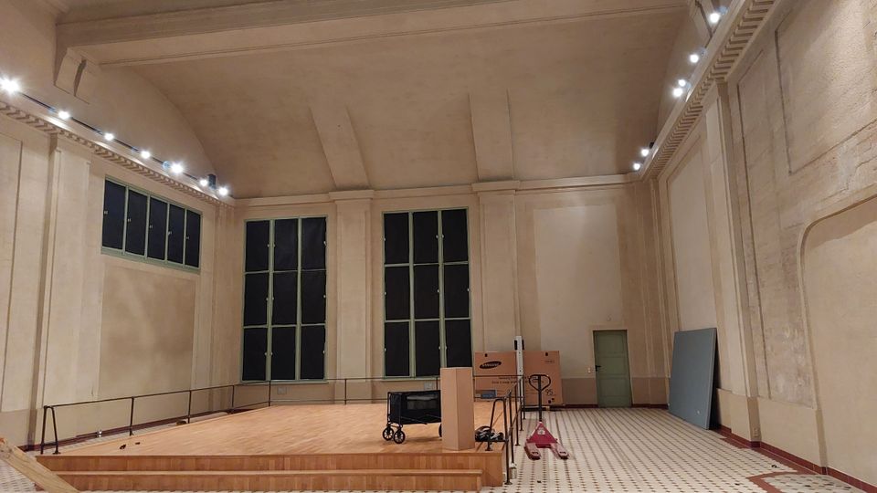 Innenraum einer großen, hellen Halle mit hoher Decke und cremefarbenen Wänden. Der Raum zeigt eine erhöhte Holzplattform mit Geländer, hohe Fenster mit grünen Rahmen, sowie einige gelagerte Gegenstände wie Kartons und Geräte; der Boden ist mit einem geometrischen Fliesenmuster gestaltet.