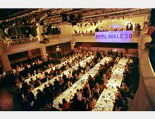 Ein großer Veranstaltungsraum mit gedeckten Tafeln, an denen zahlreiche Personen sitzen. Im oberen Bereich befinden sich Emporen, und ein beleuchtetes Banner mit der Aufschrift "BERLINALE 50" hängt an der Wand, während die Decke von Scheinwerfern und Bühnentechnik geprägt ist.