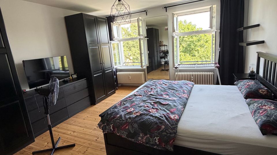 Helles Schlafzimmer mit Holzdielenboden, großem Bett und schwarzen Möbeln wie Kleiderschrank und Kommode mit Fernseher. Zwei große Fenster mit dunklen Vorhängen und Heizkörpern darunter lassen viel Tageslicht herein; an der Decke hängt eine geometrische Pendelleuchte.