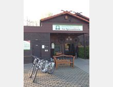 Holzgebäude mit dem Schild „Hotel & Restaurant Zur Spreewälderin“ über dem Eingang, davor stehen mehrere Elektro-Fahrräder auf einem gepflasterten Platz sowie eine hölzerne Sitzbank; flankiert von Blumen und einer Tafel mit Speiseangebot.