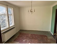 Ein leerer Raum mit hellen Wänden, einem großen Doppelfenster mit dekorativen Metallgittern, einem Heizkörper darunter sowie einem alten, abgenutzten Holzfußboden. Ein kleiner Kronleuchter hängt an der Decke, und eine Wand ist teilweise grün gestrichen.