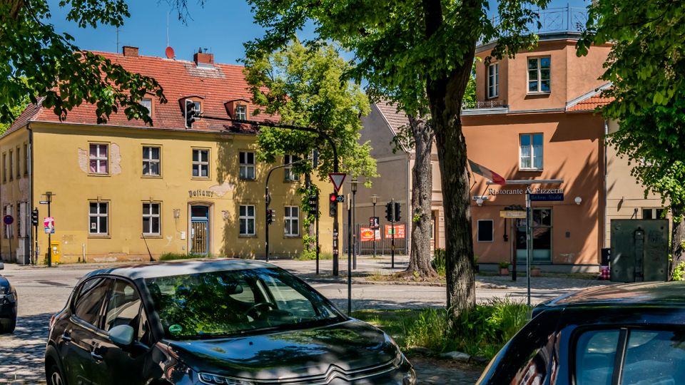 Straßenecke in einer kleinen Stadt mit gepflasterter Fahrbahn, geparkten Autos und großen Straßenbäumen im Vordergrund. Im Hintergrund stehen zweigeschossige Altbauten mit roten Ziegeldächern und hellen Fassaden, darunter ein Gebäude mit der Aufschrift „Ristorante Pizzeria“, sowie Ampeln und Verkehrszeichen an der Kreuzung.
