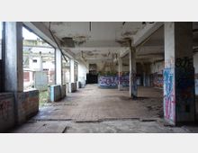 Verlassene Industriehalle mit beschädigtem Boden und teilweise eingestürzter Decke. Wände und Säulen sind mit Graffiti bedeckt, und durch die Fenster ist eine verfallene Außenstruktur sichtbar.