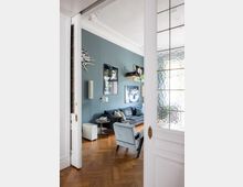 Wohnzimmer mit einer petrolblauen Wand, einem dunkelgrauen Sofa, einem hellblauen Sessel und einem Holzboden im Fischgrätmuster. Im Vordergrund sind weiße doppelte Flügeltüren mit dekorativen Verzierungen und teils geriffeltem Glaseinsatz zu sehen.