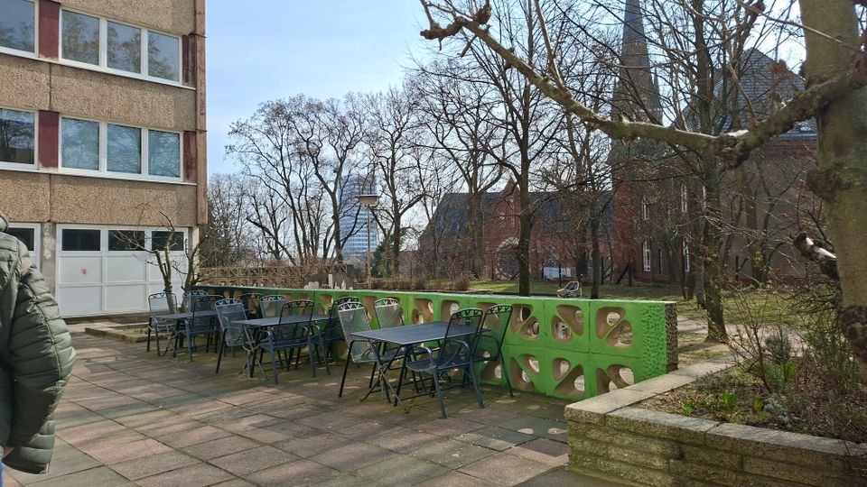 Gepflasterte Außenterrasse neben einem mehrstöckigen Gebäude mit mehreren Metalltischen und -stühlen sowie einer niedrigen grünen Betonbrüstung mit ovalen Öffnungen. Dahinter liegen ein kleiner Grünbereich mit kahlen Bäumen und im Hintergrund ein rotes Backsteingebäude mit Kirchturm.