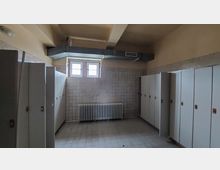 Ein Umkleideraum mit weißen Fliesenwänden, einer Reihe von weißen Spinden auf beiden Seiten, einem Heizkörper unter einem kleinen Fenster und sichtbaren Belüftungsrohren an der Decke. Der Boden besteht aus grauen Fliesen.