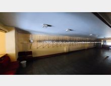 Ein leerer Garderobenbereich mit einer langen Wand aus Metallkleiderhaken und darunter verlaufenden Halterungen. Der Raum hat eine niedrige Decke, eine gelbliche Wandfarbe, dunklen Bodenbelag und vereinzelte Sitzbänke.