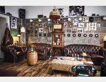 Gemütlicher Raum im Vintage-Stil mit braunen Ledersofas, einer Wanduhr, dekorativen Schallplatten und Fotografien an den Wänden. Ein Holzfußboden, ein antiker Schrank mit Glasfront und eine hölzerne Fass-Tischdekoration ergänzen die Einrichtung.