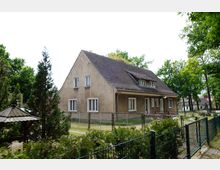 Ein zweistöckiges Wohnhaus mit hellbrauner Fassade und Schrägdach, umgeben von einem Garten mit Zaun. Im Vordergrund stehen Büsche, ein kleiner Pavillon und mehrere Bäume.