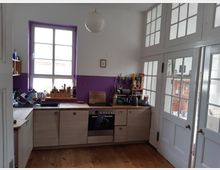 Kleine Küche mit einer Holzarbeitsplatte, weißen Schränken und einem großen Fenster mit lila Rahmen. Die Wand ist teils weiß, teils lila gestrichen, und rechts befinden sich Türen mit großen Glaseinsätzen.