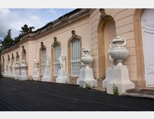 Außenansicht eines historischen Gebäudes mit beigen Wänden und großen verzierten Fenstern, flankiert von weißen Marmorskulpturen und barocken Vasen. Der Boden davor ist mit schwarzen Platten bedeckt, und im Hintergrund sind Bäume sichtbar.