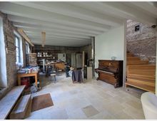 Ein großzügiger Raum mit sichtbaren Ziegelwänden, Holzbalkendecke und einer hellen Steinboden. Links stehen Arbeitstische und Regalflächen mit Materialien, rechts befindet sich ein Klavier und eine Holztreppe, die nach oben führt.