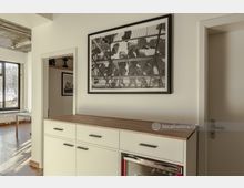 Moderne Innenansicht einer Wohnung mit einer weißen Kommode und einer Holzplatte, darüber hängt ein gerahmtes Schwarz-Weiß-Foto. Im Hintergrund sind große Fenster, eine offene Tür und ein heller Raum mit Holzboden zu sehen.