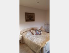 Schlafzimmer mit einem cremefarbenen Bett im klassischen Stil, dekoriert mit einer bestickten Decke und bunten Kissen. Im Hintergrund steht ein Nachttisch, und an der Wand hängt ein Bild mit Blumenmotiv.