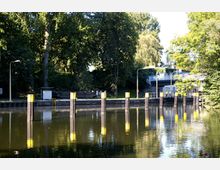 Flusslandschaft mit einer Schleuse, umgeben von dichtem Baumbestand. Im Vordergrund befinden sich stabile Holzpfeiler mit gelben Markierungen, während im Hintergrund eine Brücke und technische Gebäude erkennbar sind.