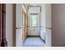 Schmaler Flur im Altbau mit hellem Holz-Eingangstür, hohen weißen Wänden mit Holzleisten und dekorativem Fliesenboden in Blau-Weiß. Am Ende befindet sich ein großes Buntglasfenster mit geometrischem Muster, daneben ein weißer Heizkörper.