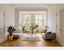 Heller Wohnraum mit hohen Decken, verzierten Stuckdetails und großen Sprossenfenstern, die auf eine grüne Außenansicht blicken. Der Raum ist mit einem Teppich, einem runden Esstisch mit Stühlen, zwei Sofas und zahlreichen Zimmerpflanzen dekoriert, der Boden ist aus Holzparkett.