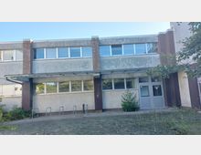 Zweistöckiges Gebäude mit grauer Fassade und großen rechteckigen Fenstern. Im Eingangsbereich befinden sich eine Überdachung, graue Türen und einige Fahrradständer, umgeben von vereinzeltem Pflanzenbewuchs.