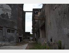 Verlassene Industrieanlage mit großen, verwitterten Betongebäuden und teils zerstörten Fenstern. Die Szenerie umfasst einen schmalen Durchgang zwischen den Gebäuden und Bewuchs am Boden sowie an den Wänden.