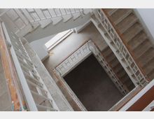 Treppenhaus mit mehreren Stockwerken, Betonstufen und weiß gestrichenen Metallgeländern. Licht fällt durch ein großes Fenster im oberen Bereich ein, und die Innenwände sind schlicht gehalten.