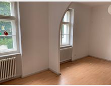 Ein leerer Raum mit hellen Wänden, Laminatboden, zwei weißen Holzfenstern mit Heizkörpern darunter und einem weißen Rundbogen, der den Raum teilt. Draußen ist grünes Laub erkennbar.