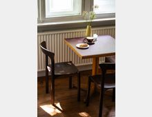 Ein kleiner Holztisch mit zwei dunklen Holzstühlen in einem hellen Raum mit Holzboden; auf dem Tisch stehen eine gelbe Vase mit Blumen, eine Kaffeetasse und ein Teller mit einem Croissant. Im Hintergrund ist ein Fenster mit einem weißen Heizkörper darunter zu sehen.