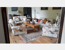 Wohnzimmer mit Holzboden, einem rechteckigen Holztisch und einem Teppich in der Mitte. Um den Tisch stehen mehrere Sessel und ein Sofa mit floralen Polstern, darauf stapeln sich Kissen und Bücher. Im Hintergrund befinden sich eine Vitrine, Lampen und an der Wand hängende Bilder.